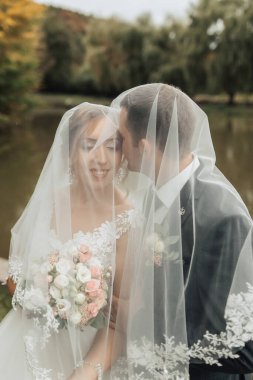 Bir gelin ve damat peçenin arkasında sarılıyorlar. Gelin elinde bir buket çiçek tutuyor.