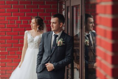 Bir adam ve bir kadın tuğla bir duvarın önünde duruyorlar. Adam takım elbise ve kravat takıyor ve kadın beyaz bir elbise giyiyor. İkisi de kameraya bakıyor.