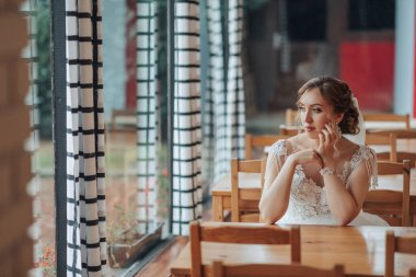 Bir gelin restoranda bir masada oturuyor, cep telefonuyla konuşuyor. Sahne ahşap sandalyeli ve pencereli bir restoranda geçiyor. Gelin bir gelinlik giyiyor ve rahatlamış.