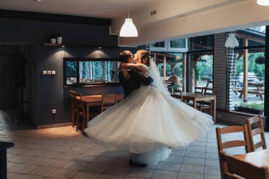 Bir gelin ve damat bir restoranda dans ediyorlar. Gelin beyaz bir elbise giyiyor ve damat bir takım elbise giyiyor. Sahne mutlu ve kutlama dolu.