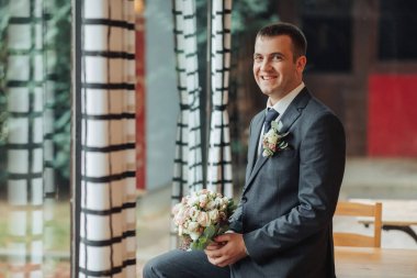 Takım elbiseli ve kravatlı bir adam gülümsüyor ve elinde bir buket çiçek tutuyor. Mutluluk ve kutlama kavramı, muhtemelen bir düğün ya da resmi bir toplantı gibi özel bir etkinliğe katılacaktır.