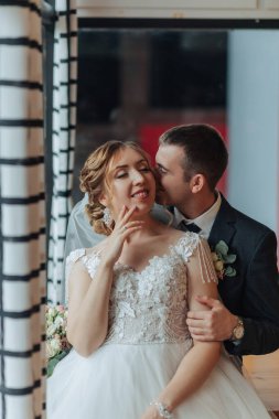 Bir gelin ve damat pencerenin önünde öpüşüyorlar. Gelin beyaz bir elbise giyiyor ve damat da takım elbise giyiyor.