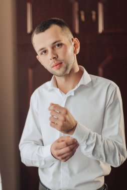 Beyaz gömlekli bir adam bir kapının önünde duruyor. Gömleğini düzeltiyor ve kameraya bakıyor.