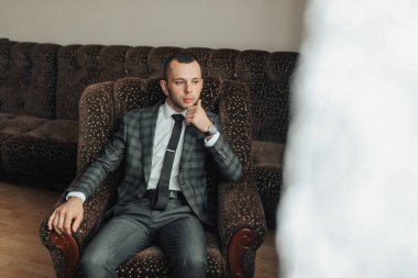 Takım elbiseli ve kravatlı bir adam sandalyede oturuyor. Bir şeye bakıyor ve derin düşüncelere dalmış.