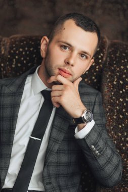 Takım elbiseli ve kravatlı bir adam sandalyede oturuyor ve aşağıya bakıyor. O bir saat ve kravat takıyor.