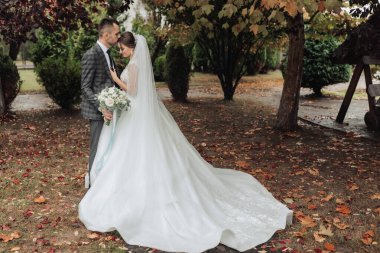 Gelin ve damat parkta duruyorlar, gelin beyaz bir elbise giyiyor ve damat elinde buket tutuyor.