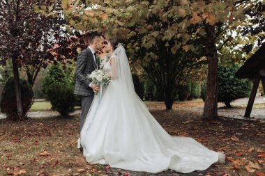 Parkta bir gelin ve damat öpüşüyor. Gelin beyaz bir elbise giyiyor ve damat da takım elbise giyiyor.