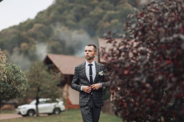 Takım elbiseli ve kravatlı bir adam ormanda yürüyor. Orman yemyeşil ve yemyeşil. Arka planda park etmiş bir araba var.