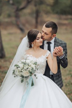 Bir gelin ve damat birbirlerinin yanaklarını öpüyor. Gelin beyaz bir elbise giyiyor ve damat da takım elbise giyiyor.