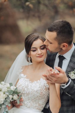 Bir gelin ve damat birbirlerinin ellerini öpüyor. Gelin beyaz bir elbise giyiyor ve damat da takım elbise giyiyor.