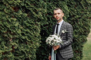 Takım elbiseli ve kravatlı bir adam elinde bir buket çiçekle çalıların önünde duruyor.