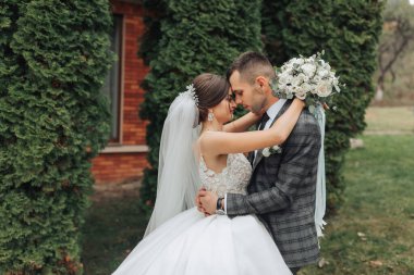 Bir gelin ve damat bir binanın önünde birbirini kucaklıyor. Gelin beyaz bir elbise giyiyor ve damat bir takım elbise giyiyor. El ele tutuşup kameraya gülümsüyorlar.