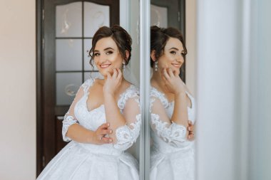 Beyaz elbiseli bir kadın aynanın önünde duruyor. Gülümsüyor ve yansımasına bakıyor.