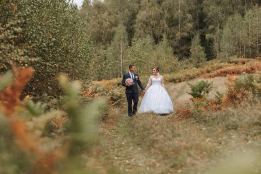 Bir gelin ve damat ormanda el ele tutuşup gülümsüyor. Sahne mutlu ve romantik.