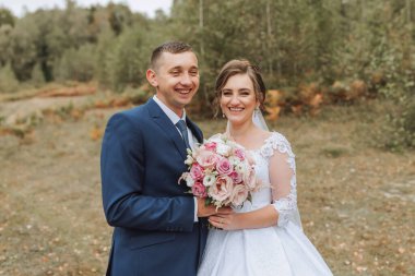 Bir gelin ve damat tarlada fotoğraf çektirmek için poz veriyorlar. Gelin elinde bir buket pembe çiçek tutuyor.