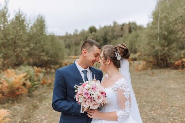 Bir gelin ve damat tarlada kameraya gülümsüyor. Gelin bir buket çiçek tutuyor ve damat kravat takıyor. Sahne mutlu ve neşeli.