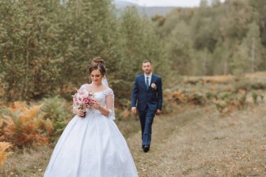 Bir gelin ve damat bir buket ve kravatla bir tarlada yürüyorlar. Gelin beyaz bir elbise giyiyor ve damat mavi bir takım elbise giyiyor. Sahne romantik ve mutlu.