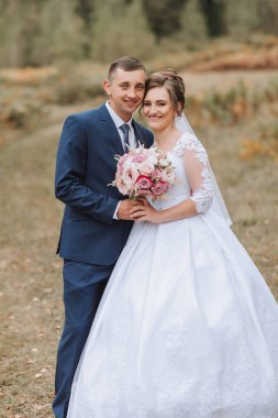 Bir gelin ve damat bir buket çiçekli fotoğraf için poz veriyorlar. Gelin beyaz bir elbise giyiyor ve damat mavi bir takım elbise giyiyor. Sahne mutlu ve kutlama dolu.