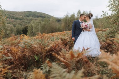 Bir gelin ve damat kahverengi çimlerin üzerinde duruyor. Gelin beyaz bir elbise giyiyor ve damat mavi bir takım elbise giyiyor. El ele tutuşup kameraya gülümsüyorlar.