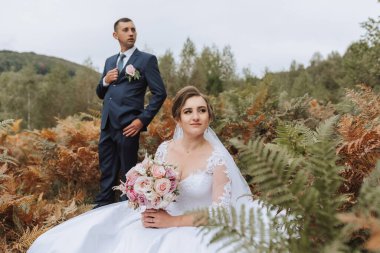 Bir gelin ve damat tarlada fotoğraf çektirmek için poz veriyorlar. Gelin bir buket çiçek tutuyor ve damat takım elbise giyiyor. Sahne mutlu ve romantik.