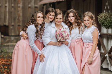 Bir gelin ve beş nedimesi fotoğraf için poz veriyor. Gelin beyaz bir elbise ve beyaz bir ceket giyiyor. Gelin elinde bir buket çiçek tutuyor.