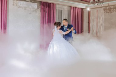 Bir gelin ve damat sisli bir odada dans ediyorlar. Gelin beyaz bir elbise giyiyor ve damat mavi bir takım giyiyor.