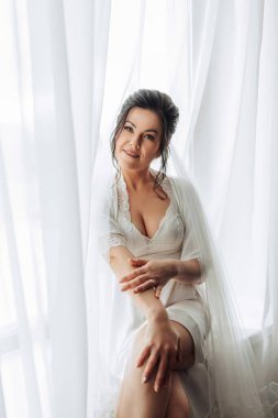 Beyaz elbiseli bir kadın perdenin önünde oturuyor. Gülümsüyor ve mutlu.