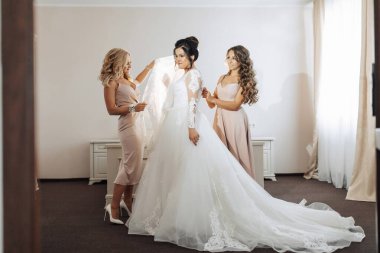 Üç kadın düğün için hazırlanıyor. İçlerinden biri uzun beyaz bir elbise giyiyor. Diğer iki kadın giyinmesine yardım ediyor.