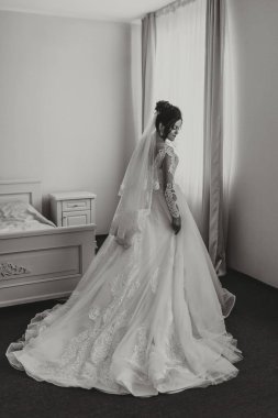 Gelinlik giymiş bir kadın yatağın önünde duruyor. Oda loş bir şekilde aydınlatılmış ve kadın bir resim için poz veriyor. Sahne zarif ve romantik.