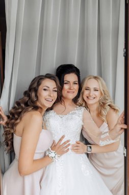 Üç kadın fotoğraf için poz veriyor, biri gelinlik giyiyor. Sahne mutlu ve kutlama dolu. Kadınlar gülümsüyor ve kameraya poz veriyor.