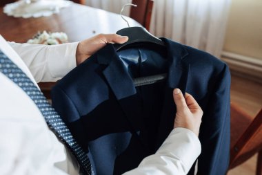 Bir adam askıda mavi bir ceket tutuyor. Ceket ahşap bir sandalyede asılı.