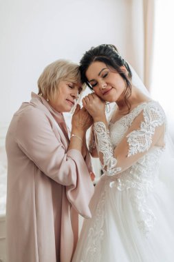 Bir kadın düğünü için hazırlanıyor ve annesi ona yardım ediyor. Gelin beyaz bir elbise giyiyor ve anne pembe bir ceket giyiyor. Sahne, iki kadın arasındaki sevgi ve destektir.