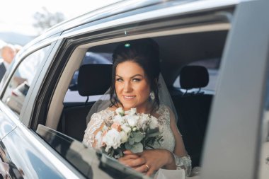 Bir gelin elinde bir buket çiçekle arabada oturuyor. Gülümsüyor ve pencereden dışarı bakıyor.