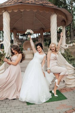 Üç kadın çardağın önünde fotoğraf çektirmek için poz veriyor. Kadınlardan biri beyaz bir elbise giyiyor ve elinde bir buket çiçek tutuyor. Diğer iki kadın da elbise giyiyor.