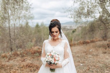 Bir gelin elinde bir buket çiçekle bir tarlada duruyor. Sahne sakin ve huzurlu, gelin uzaklara bakıyor.