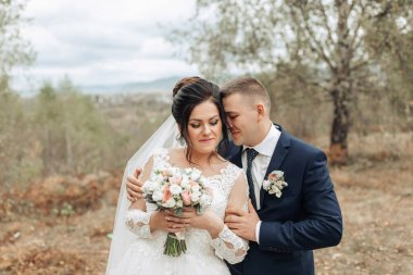 Bir gelin ve damat tarlada birbirlerine sarılıyorlar. Gelin elinde bir buket çiçek tutuyor.