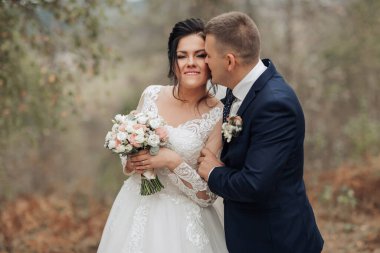 Bir gelin ve damat ormanda birbirlerine sarılıyorlar. Gelin elinde bir buket çiçek tutuyor.