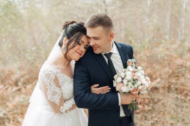 Bir gelin ve damat ormanda birbirlerine sarılıyorlar. Gelin elinde bir buket çiçek tutuyor.