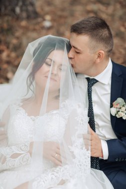 Bir gelin ve damat bir peçenin altında birlikte oturuyorlar. Gelin beyaz bir elbise giyiyor ve damat mavi bir takım elbise giyiyor. Birbirlerini öpüyorlar ve gelin peçeyle örtülü.