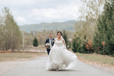 Bir gelin ve damat uzun beyaz bir elbise giymiş gelinle yolda yürüyorlar. Sahne arka planda ağaçların olduğu kırsal bir alanda geçiyor. Sahne romantik ve mutlu.