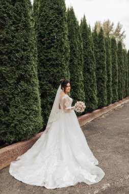 Beyaz elbiseli bir kadın çitin önünde duruyor. Elinde bir buket çiçek tutuyor.