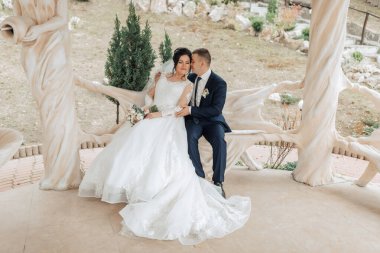 Gelin ve damat bankta oturuyor, gelin beyaz bir elbise ve damat mavi bir takım elbise giyiyor.