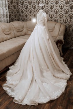 Uzun beyaz bir elbise askıda asılı. Kanepe kahverengi ve üzerinde bir desen var. Görüntü zarafet ve entelektüellik hissi veriyor.