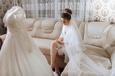 Bir gelin kafasında beyaz bir peçeyle bir kanepede oturuyor. Yanındaki elbiseye bakıyor.