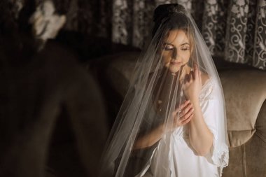 Peçeli bir kadın kanepede oturuyor. Beyaz bir elbise ve duvak takıyor.