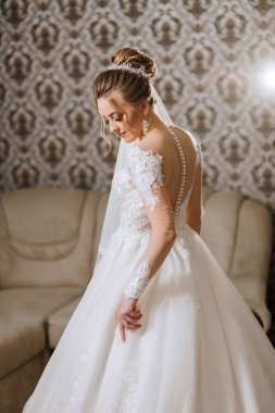 Gelinlikli bir kadın kanepenin önünde duruyor. Duvak ve kolye takıyor.