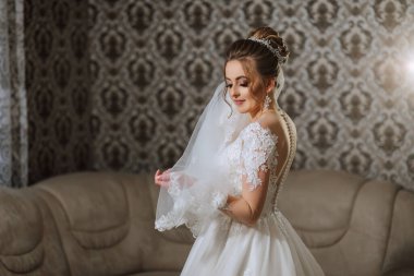 Gelinlikli bir kadın kanepenin önünde duruyor. Elinde bir peçe tutuyor.