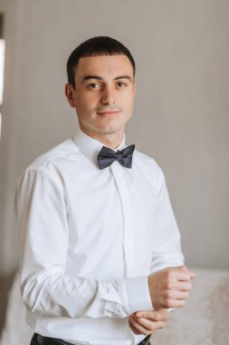 Beyaz gömlekli ve mavi papyonlu bir adam bir duvarın önünde duruyor. Kravat takıyor ve elleri cebinde.