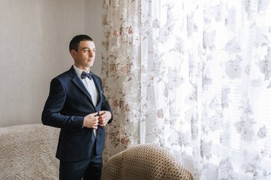 Takım elbiseli bir adam dışarı çıkmaya hazırlanıyor. O bir kravat takıyor ve ceketini ayarlıyor. Odada çiçek perdesi ve sandalye var.