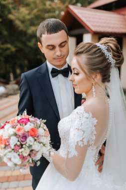 Gelin ve damat birlikte duruyorlar, gelin elinde bir buket çiçek tutuyor. Gelin beyaz bir elbise giyiyor ve damat bir takım elbise giyiyor. Sahne mutlu ve kutlama dolu.